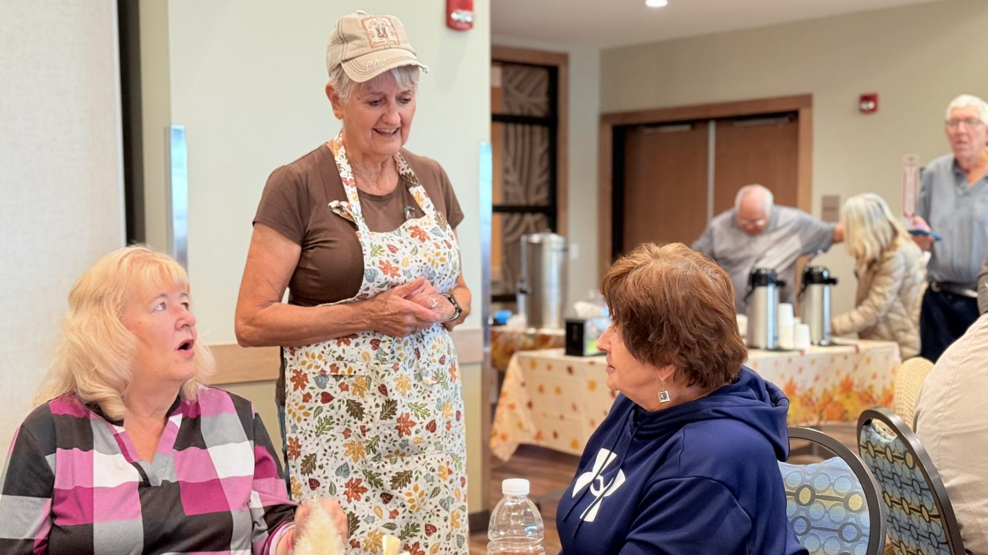One older woman in an apron talking with two older women seated at a table enjoying food and beverages.